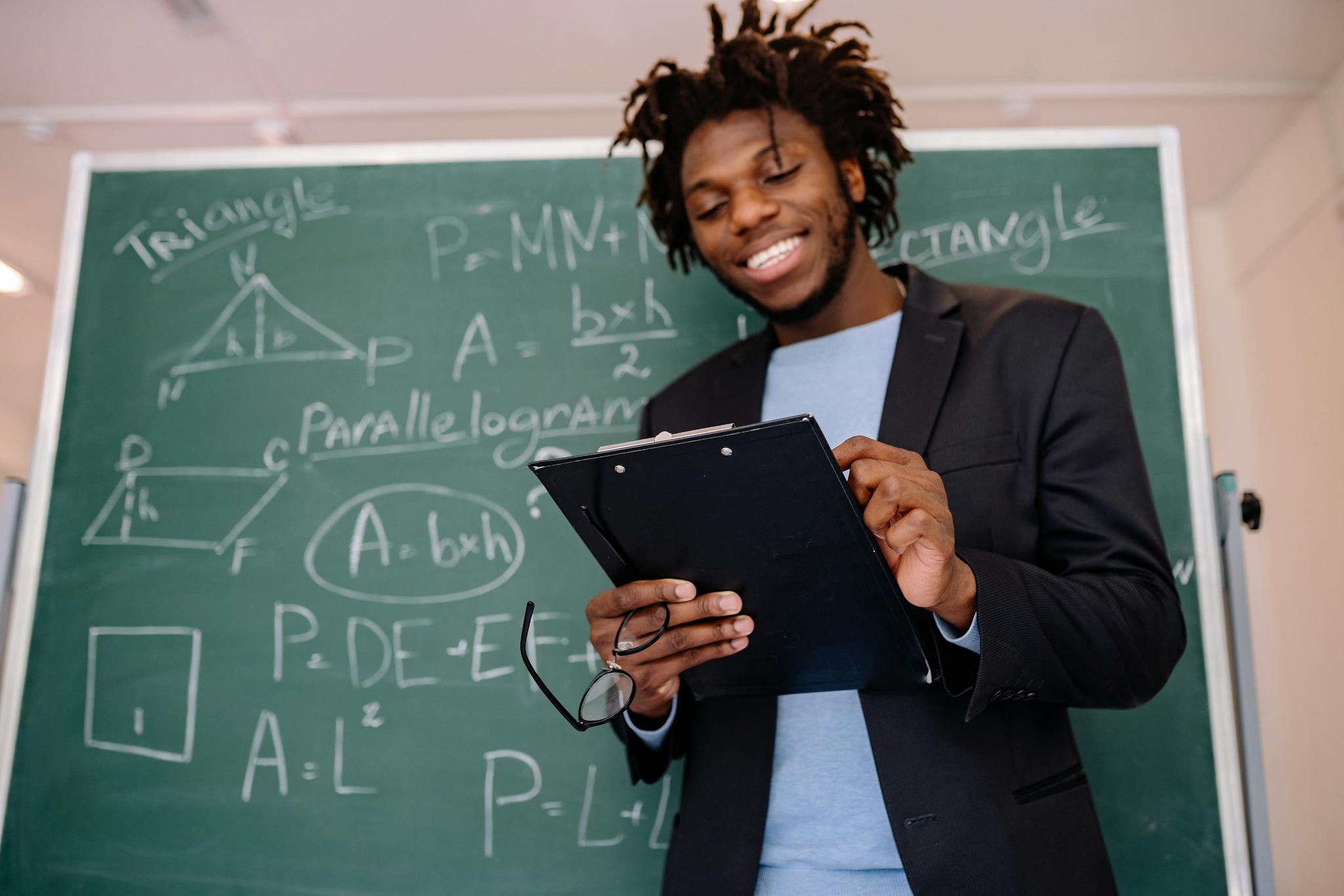 A happy teacher standing in front of a blackboard with geometric formulas, holding a clipboard.