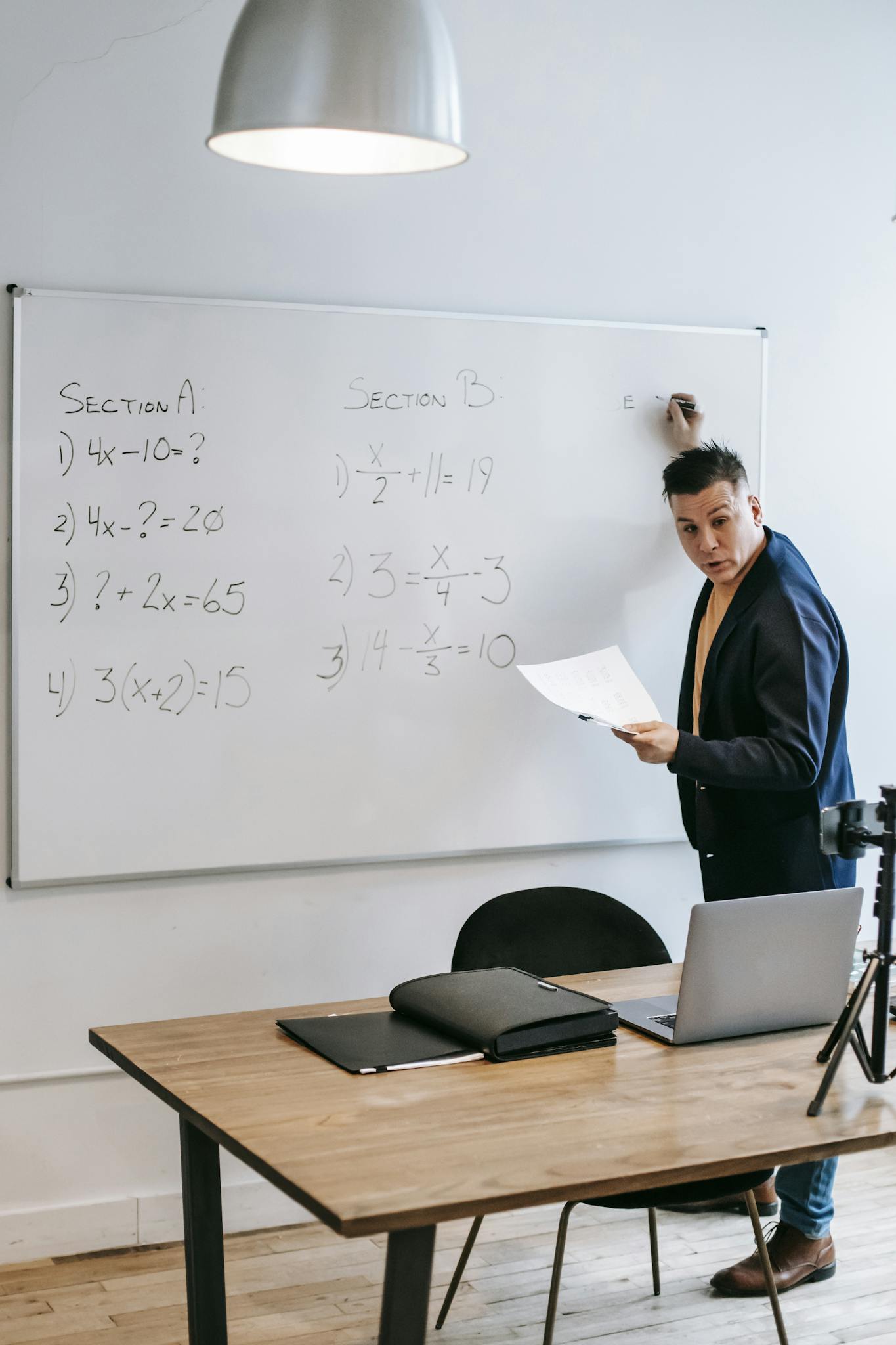 Math instructor teaches equations on a whiteboard using a laptop and tripod setup.